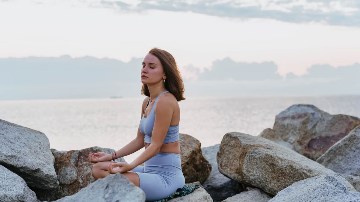 Woman in activewear meditating peacefully on rocks by the ocean at sunrise, promoting wellness and tranquility.